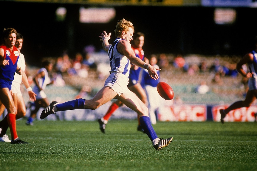 A VFL player wearing blue and white in the action of kicking the ball downfield as opponents watch during a game. 