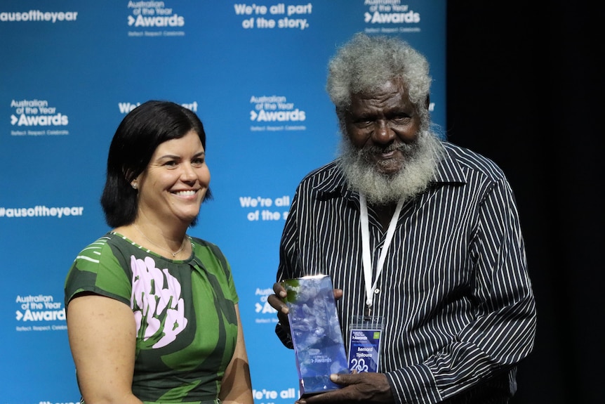 an elderly aboriginal man wearing a stripey collared top next to a woman wearing a green dress