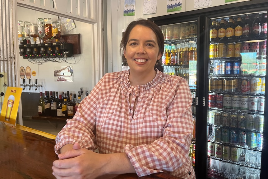 A woman in red and white checks smiles while leaning on a bar, in front of a fridge full of beers.