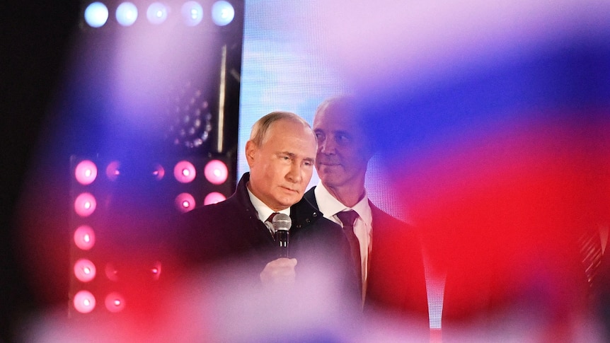 Putin with an impassive look on his face, surrounded by Russian flags in soft focus