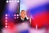 Putin with an impassive look on his face, surrounded by Russian flags in soft focus