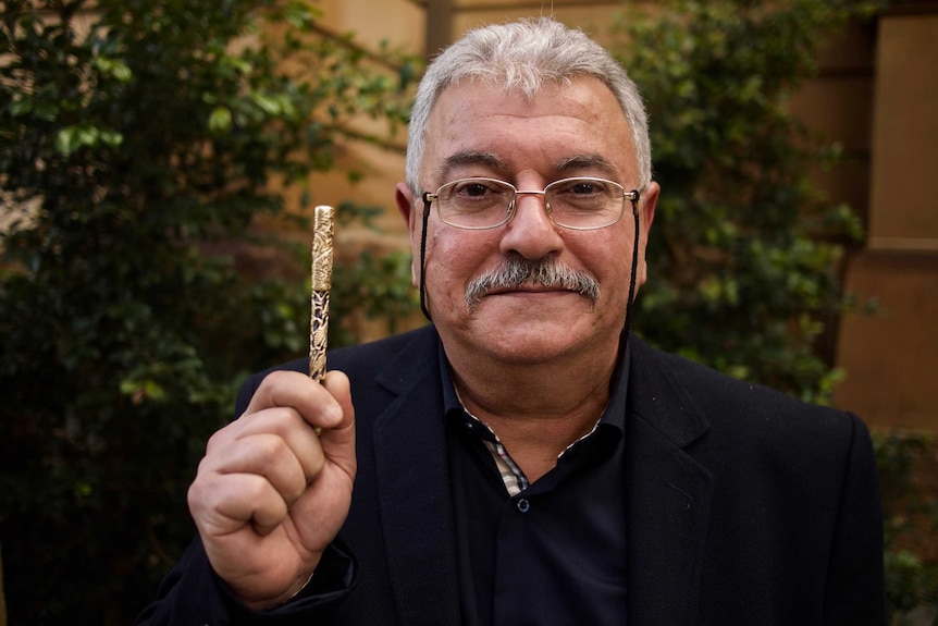 Sameer Dakhil dressed in a navy jacket and shirt holds a golden pen he carved.