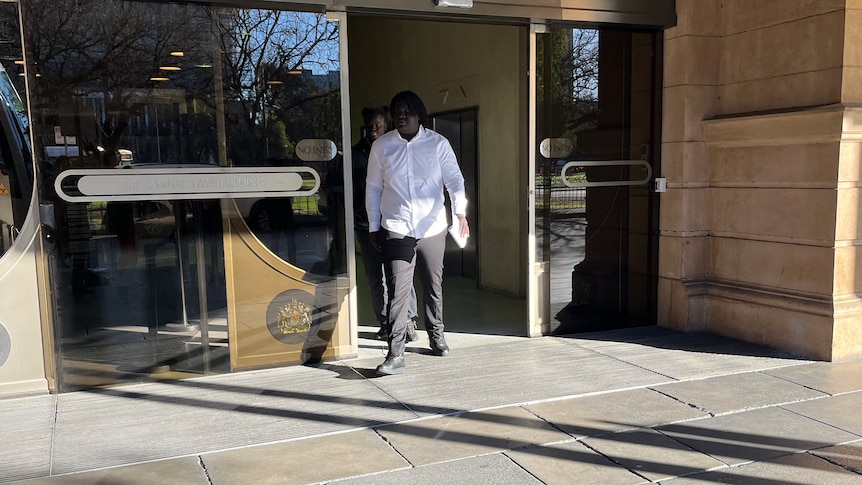 A man dressed in a white shirt and black pants walks out from glass doors.