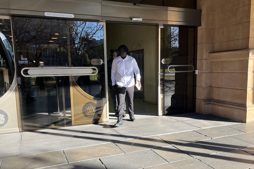 A man dressed in a white shirt and black pants walks out from glass doors.
