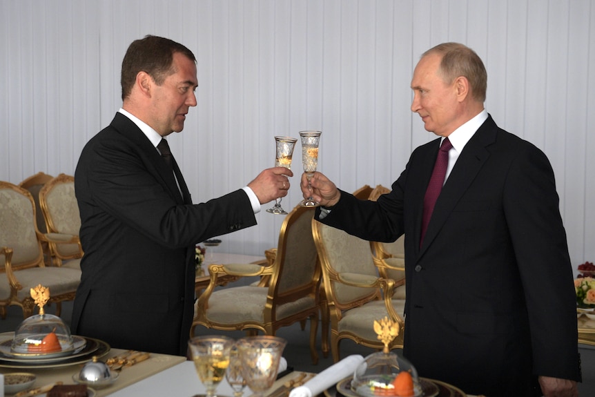 Vladimir Putin and Dimitri Medvedev toast each other with gold champagne flutes, both men are smiling 