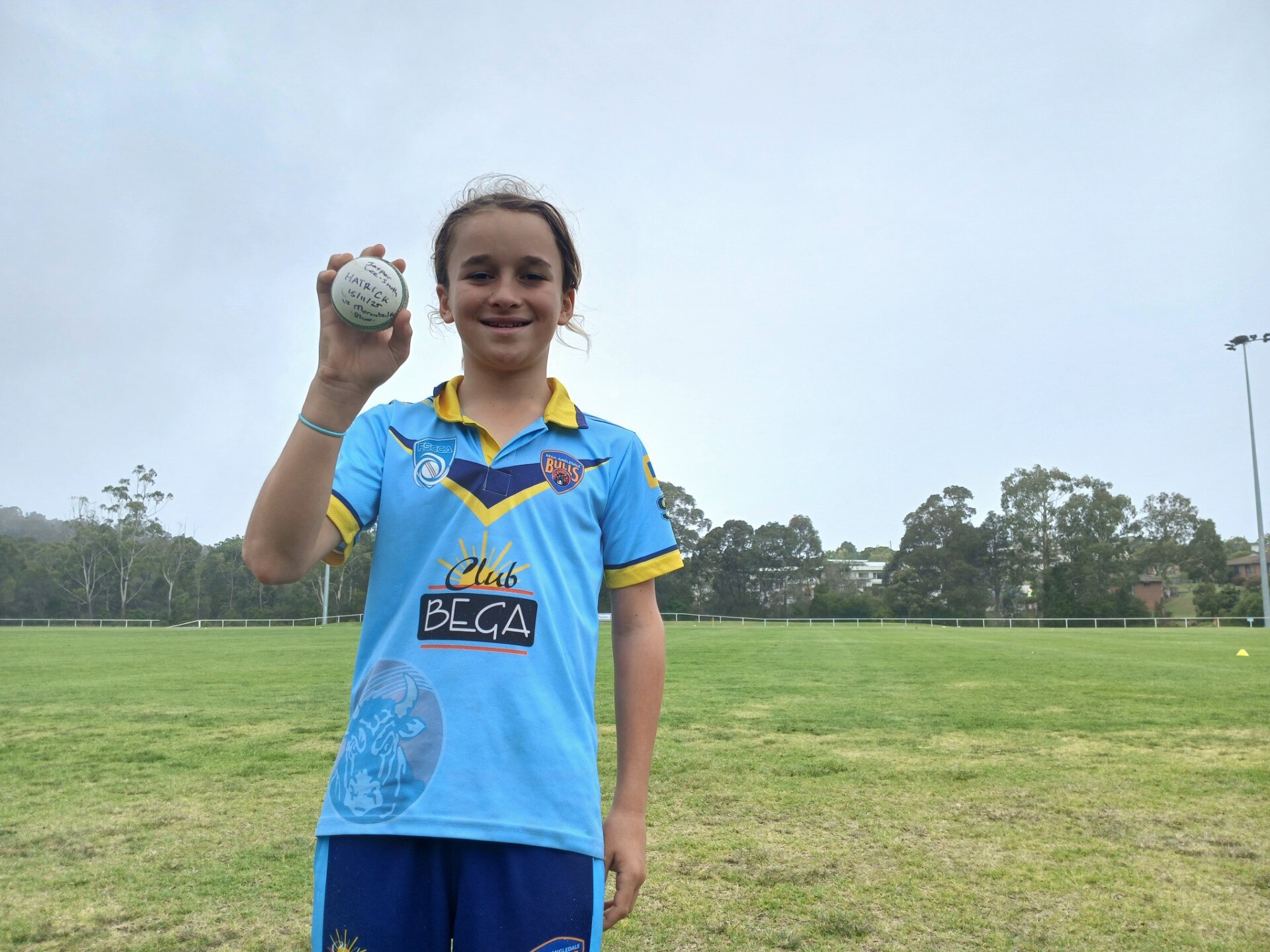 A boy in a cricket uniform smiles and holds up a cricket ball.