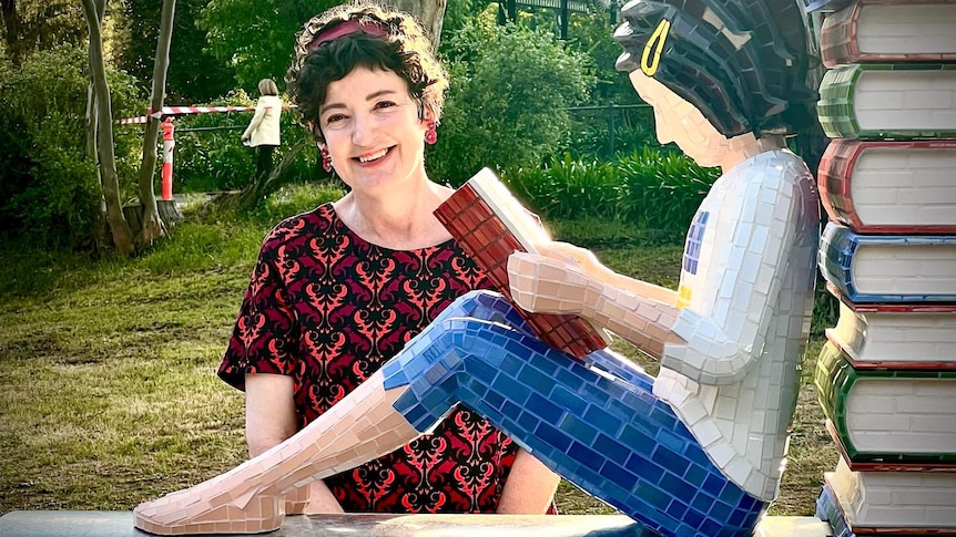 A smiling woman with curly dark hair stands behind a statue of a child sitting on a roof and reading a book.