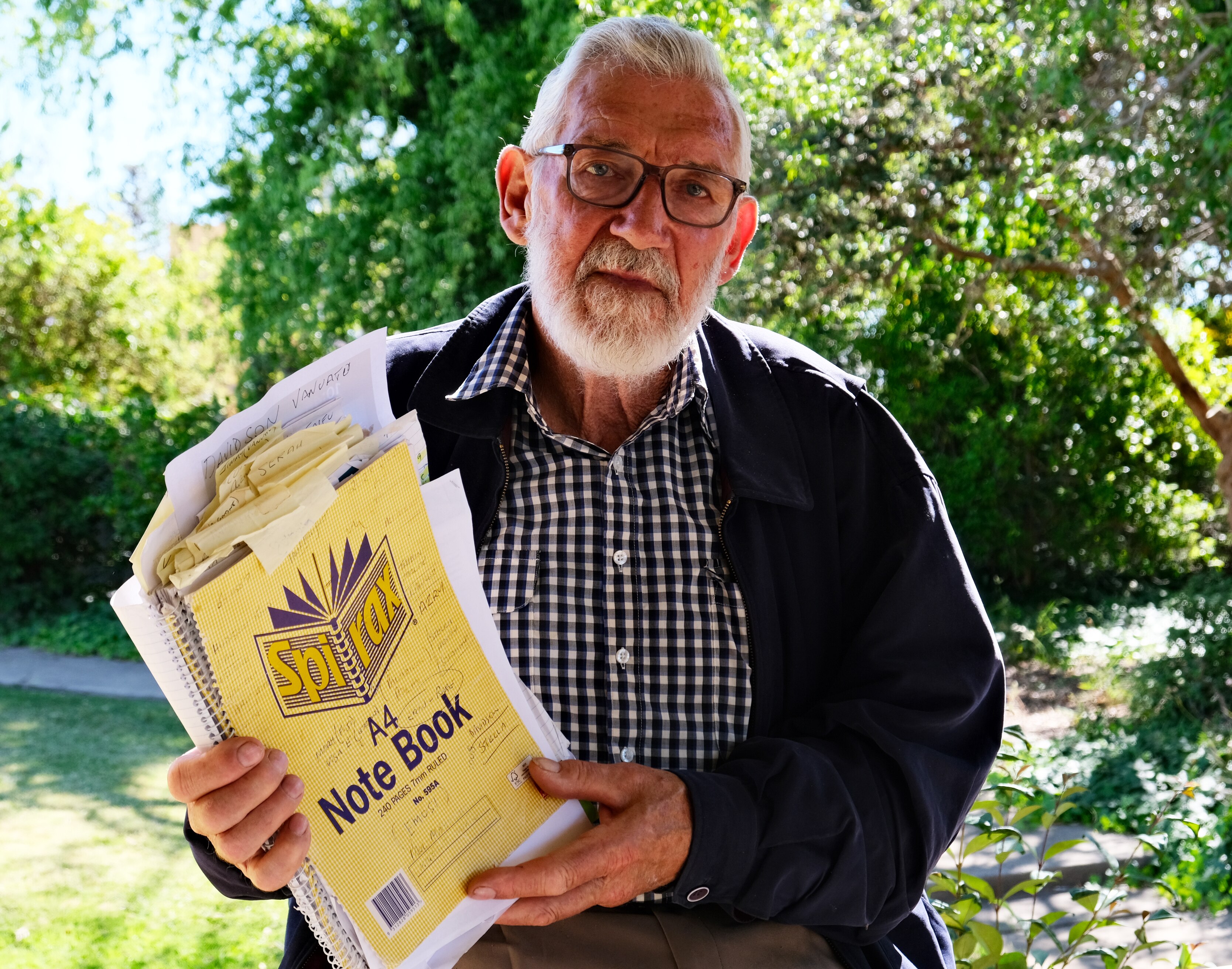 A man wearing a checked shirt and blue jacket. He has a grey beard and is wearing glasses and holding a notepad with loose paper