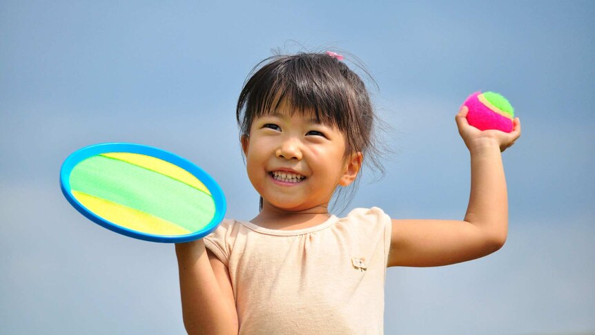 A young girl is about to throw a green-and-pink ball with her left hand. She is holding a flat disc in her right hand.
