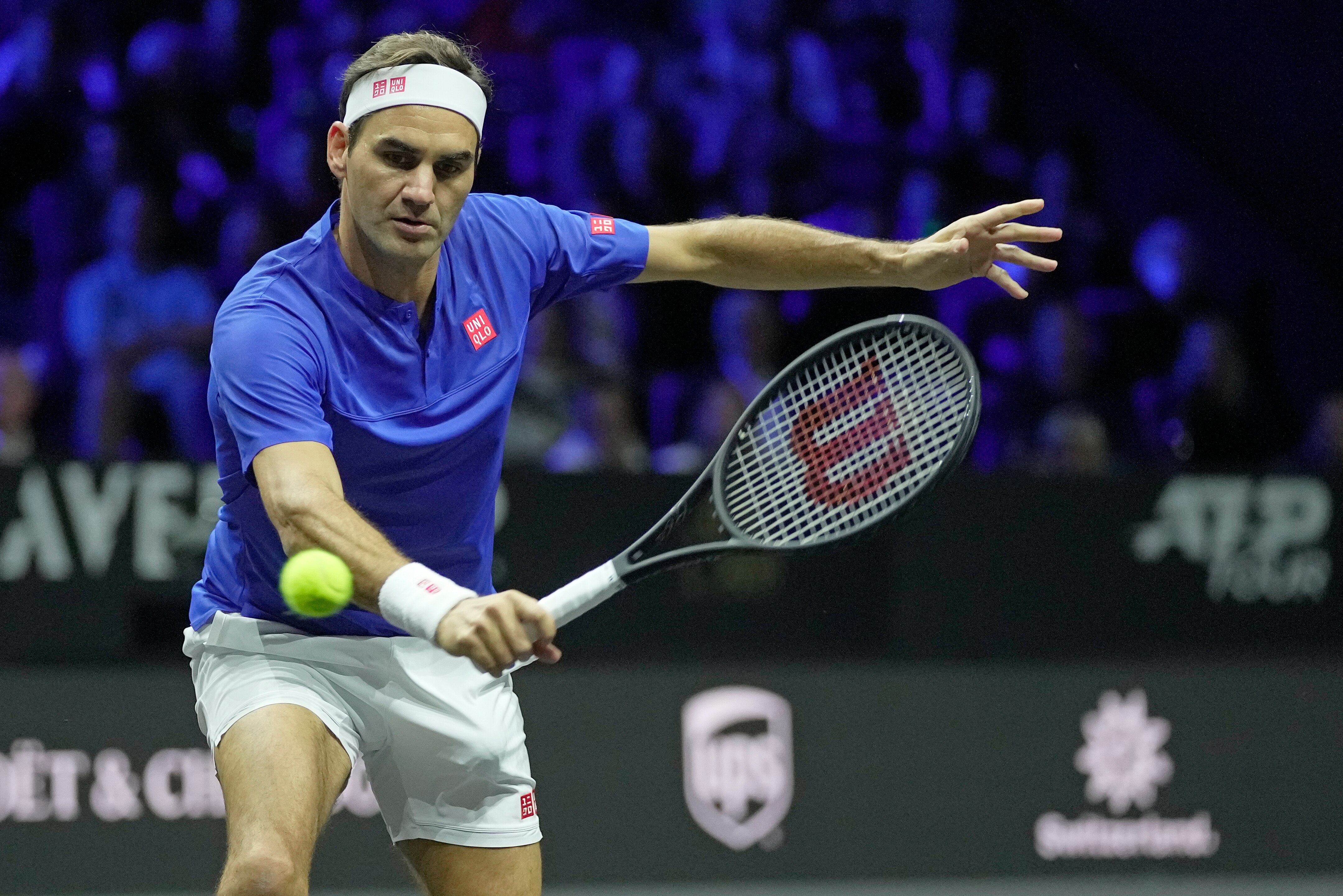 Rodger Federer in a blue shirt and white shorts leaning forward with racket in hand to take a shot on the tennis court