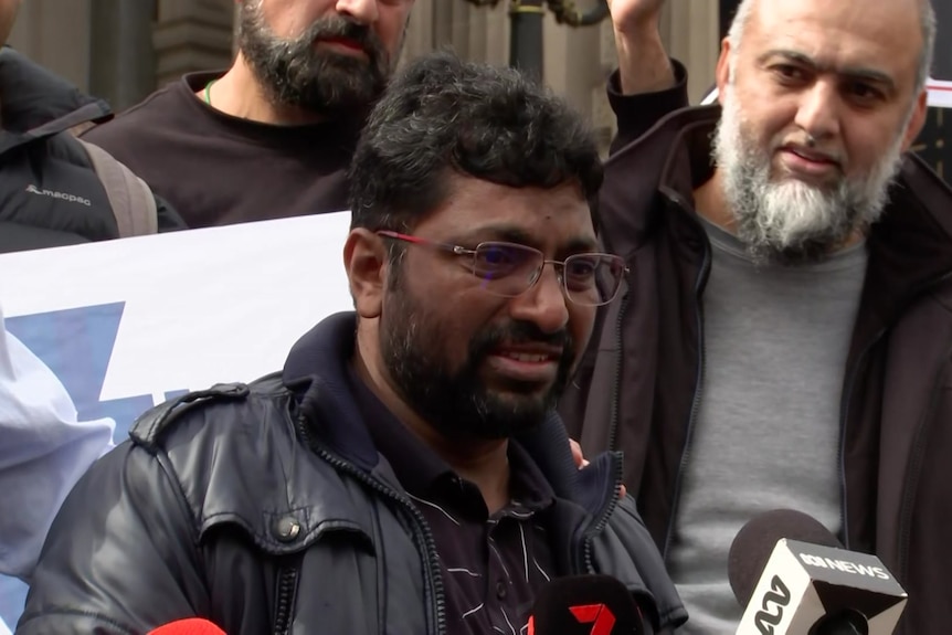 A man wearing glasses and a black jacket shows emotion as he speaks into a microphone.