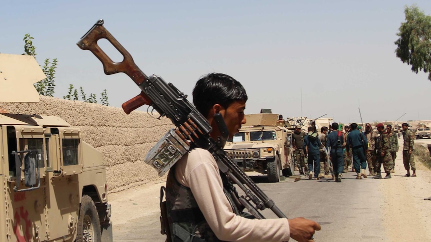 An Afghan man preparing for combat, holding a large gun surrounded by military vehicles and soldiers.