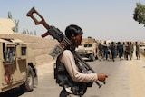 An Afghan man preparing for combat, holding a large gun surrounded by military vehicles and soldiers.