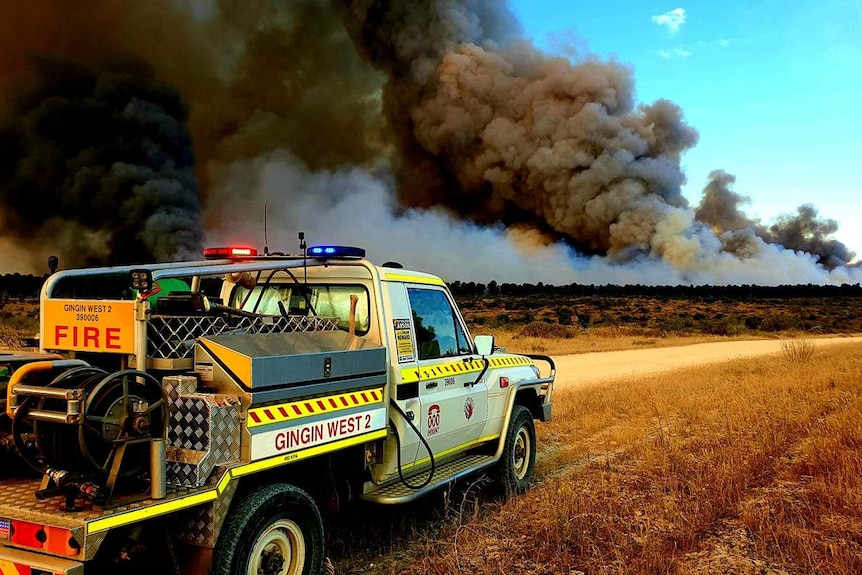 A small fire truck is parked near a sandy track with a large plume of black smoke in the background.