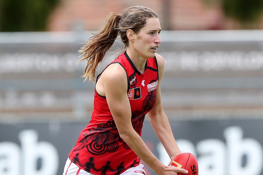 A woman Aussie rules player wearing red and white runs with the ball in her hands