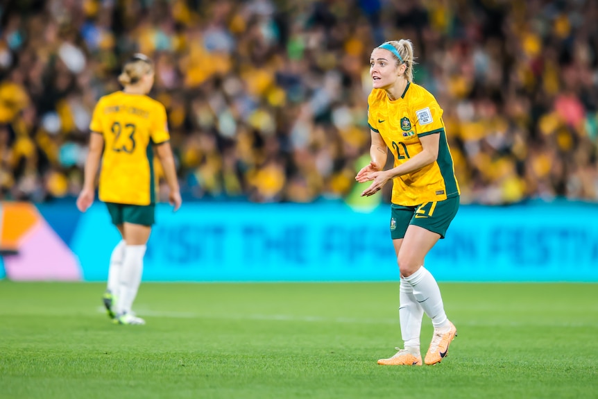 A soccer player wearing yellow and green claps during a game with a team-mate and a crowd behind her
