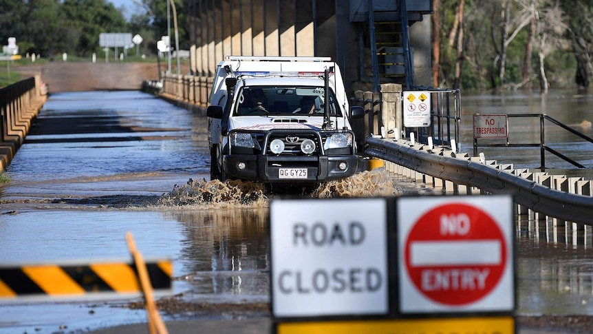 A four-wheel drive ute drives over a partially flooded river bridge