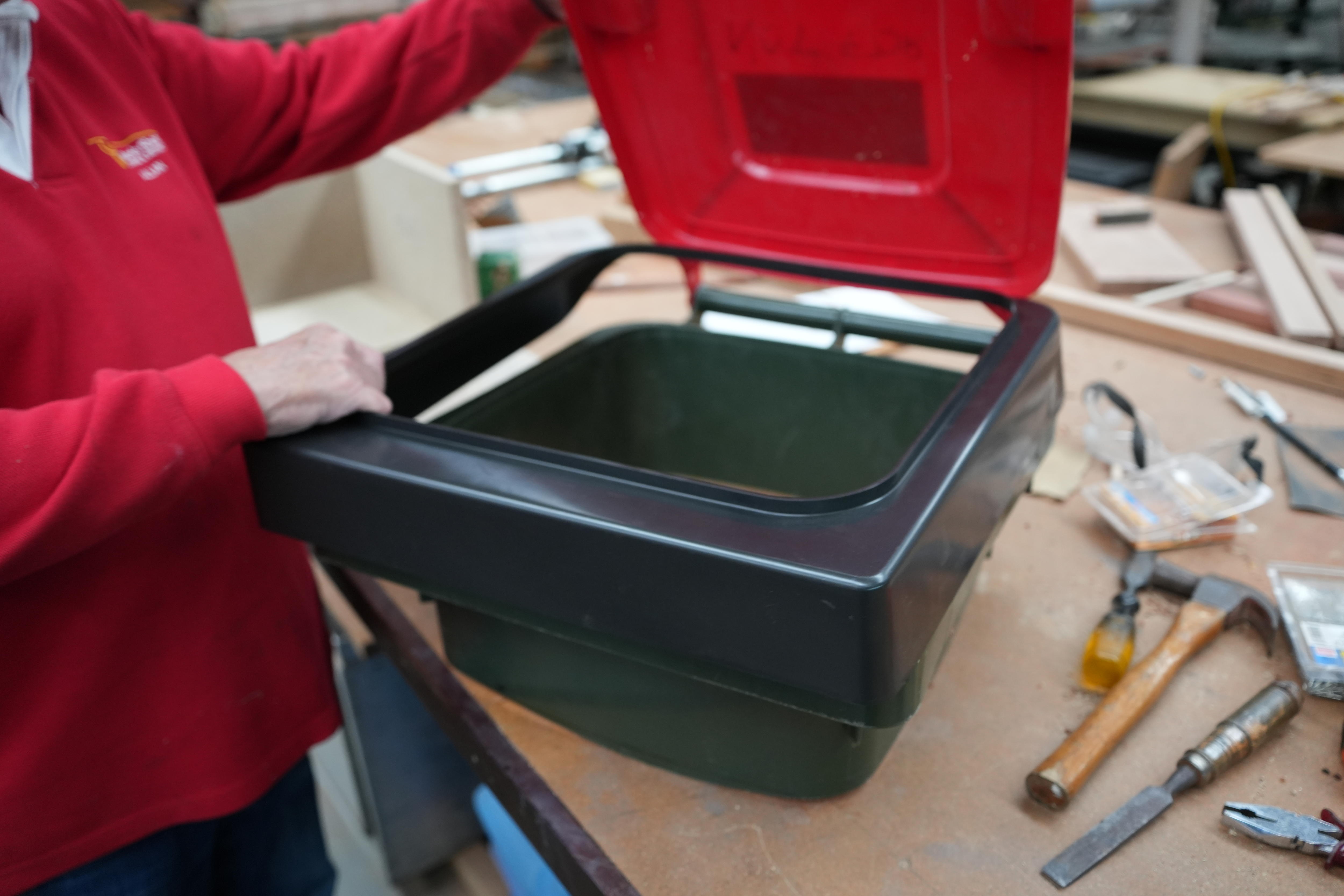 A person in a red shirt holds open the lid of a wheelie bin which has a dark plastic square fitted inside.