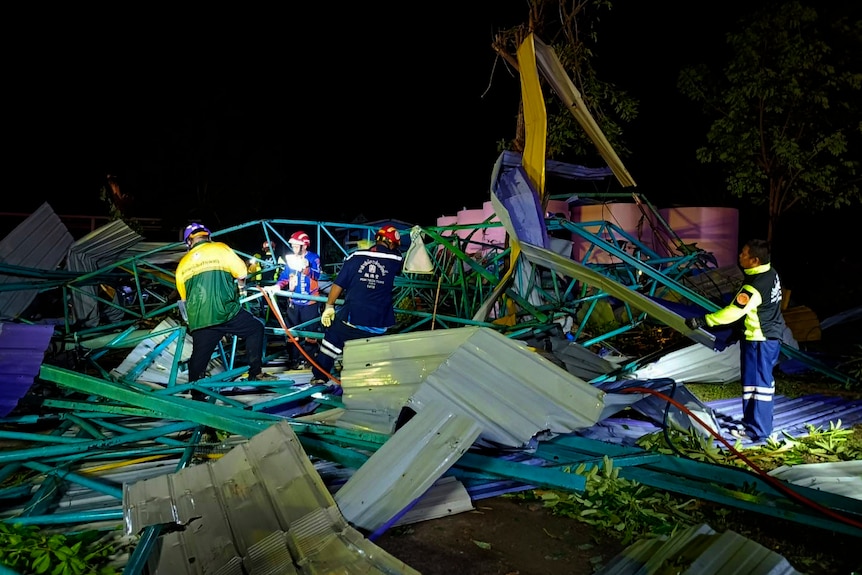 Rescue workers toil under lights at the scene of a collapsed metal roof, with colourful twists of metal wreckage across ground.
