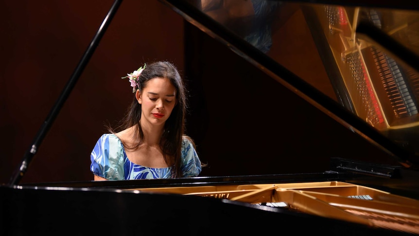 A photo of Mahani Teave through a grand piano. She has a flower in her hair, a blue and white floral dress and is concentrating.