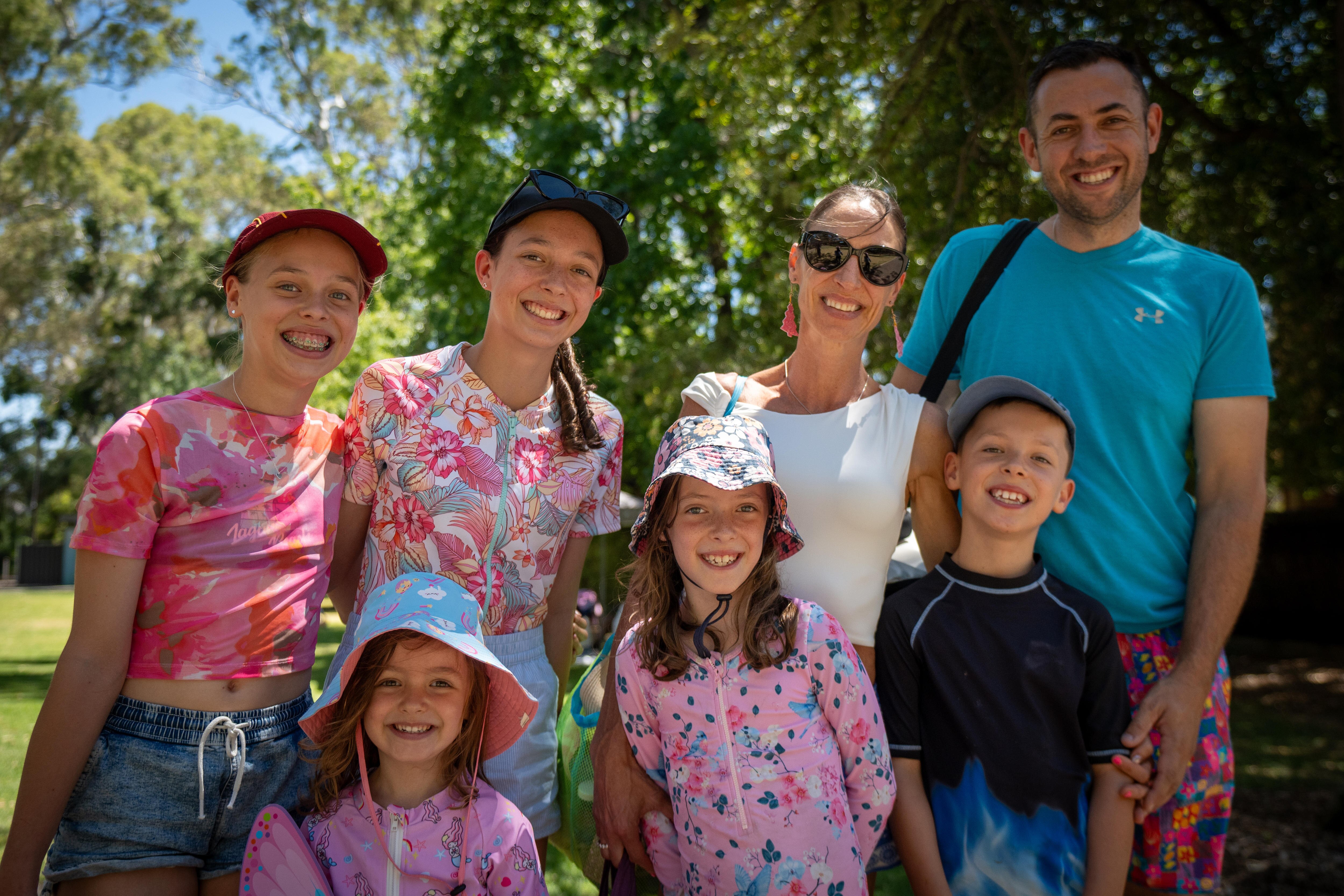 Family stands together smiling dressed in colourful clothing, leafy trees in background