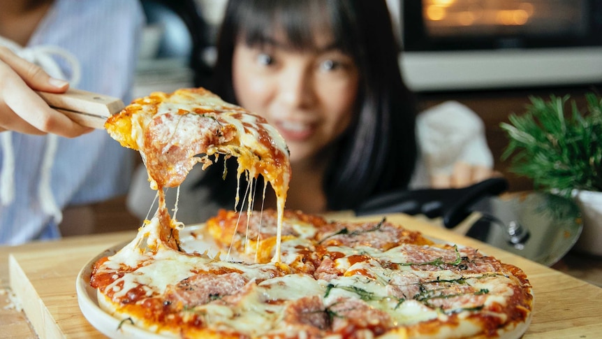A hot oozy, cheesey pizza sits in the foreground, once slice held up with a good cheese pull. A woman look on in the background.