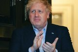 Boris Johnson, a pale man with blonde hair, claps his hands while standing in front of an open door.