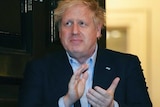Boris Johnson, a pale man with blonde hair, claps his hands while standing in front of an open door.