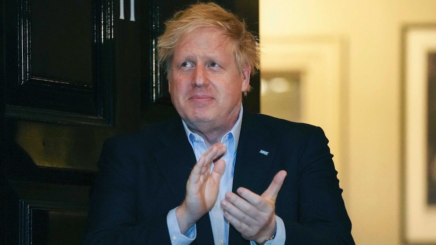 Boris Johnson, a pale man with blonde hair, claps his hands while standing in front of an open door.