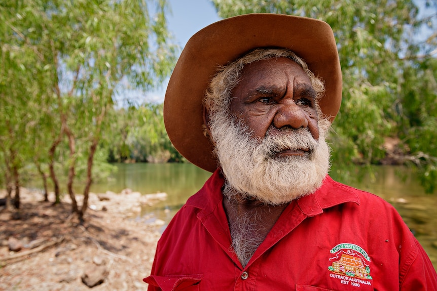 An Indigenous man on the banks of a river in Borroloola in a red shirt and hat.