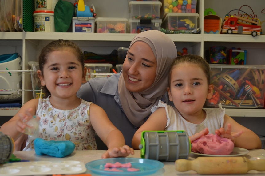 Two children with their mother standing in between them sit at a desk with play dough