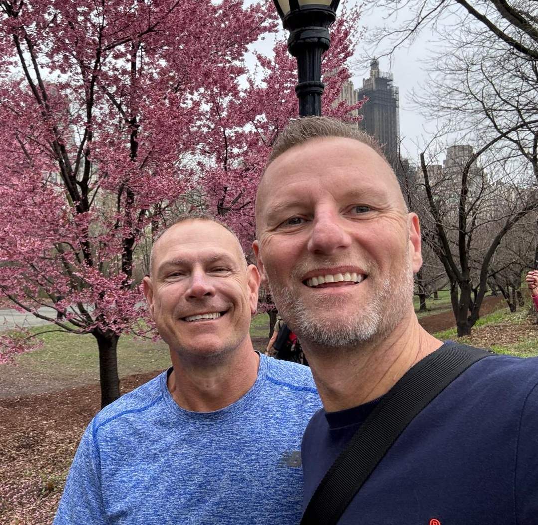 Two men in blue shirts are standing in a garden and smiling at the camera