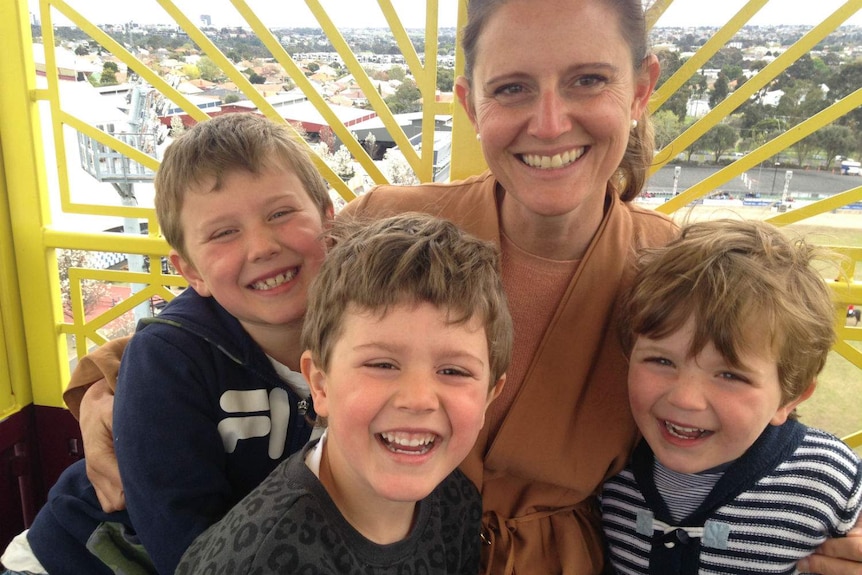 A smiling woman has her arms around three smiling young boys.