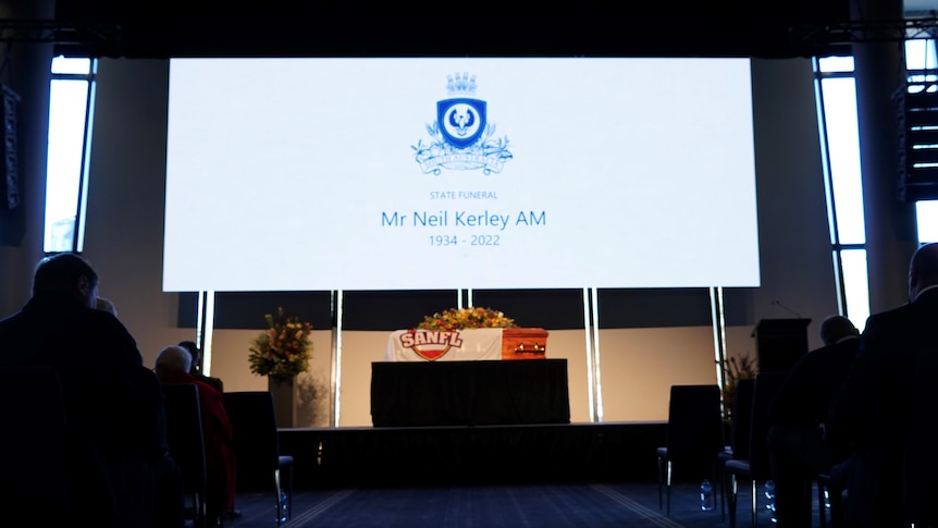 a coffin with flowers on stage with the screen showing the SA logo, Mr Neil kerley AM 