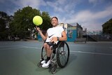 Wheelchair tennis star Dylan Alcott smiles as he is pictured in a street park holding an oversize tennis ball.