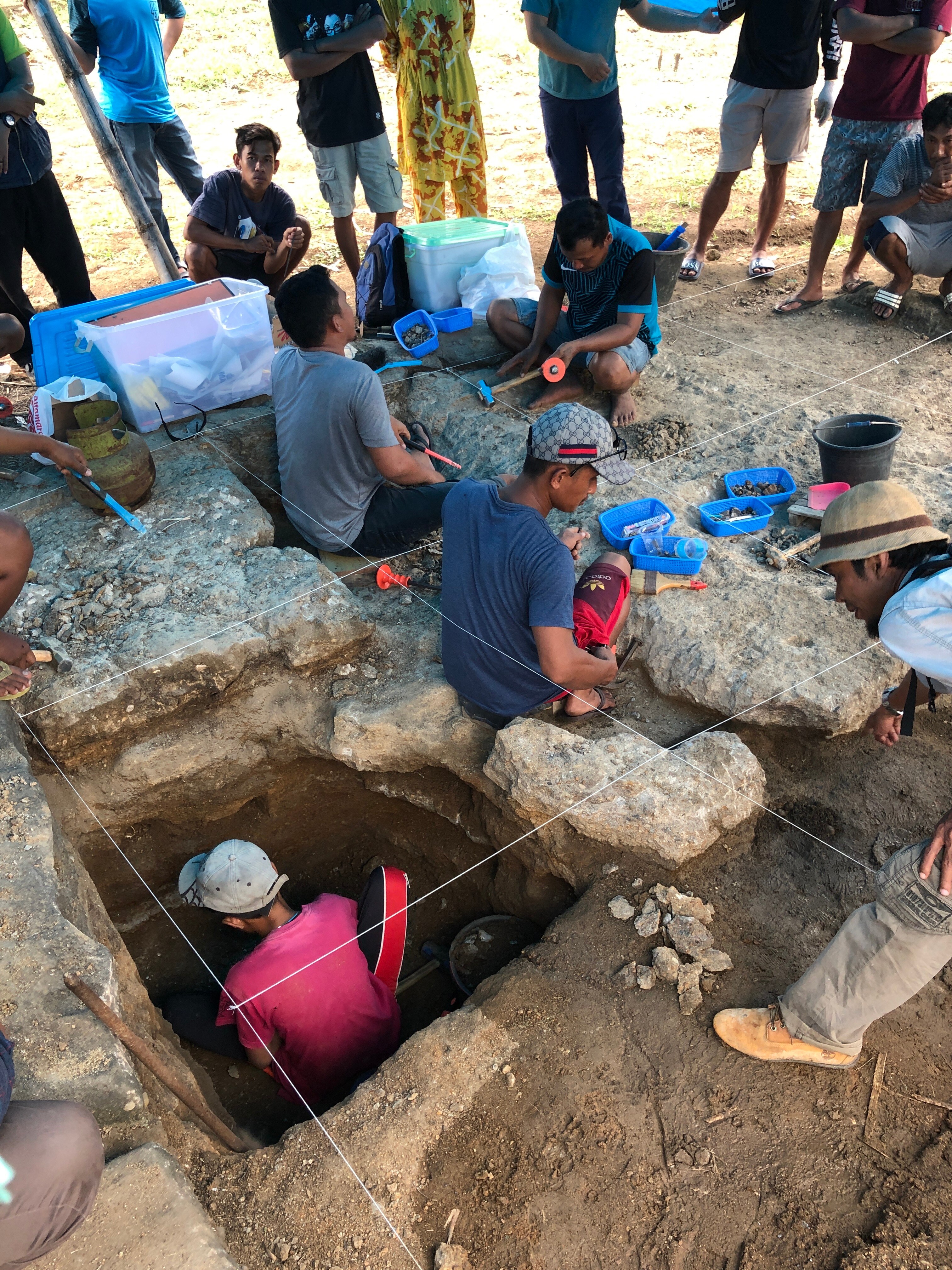 One person standing in a square rocky hole surrounded by other people in caps and tshirts crouching and standing.