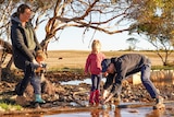 A man, woman and two children watch as the man fixes a hose outdoors