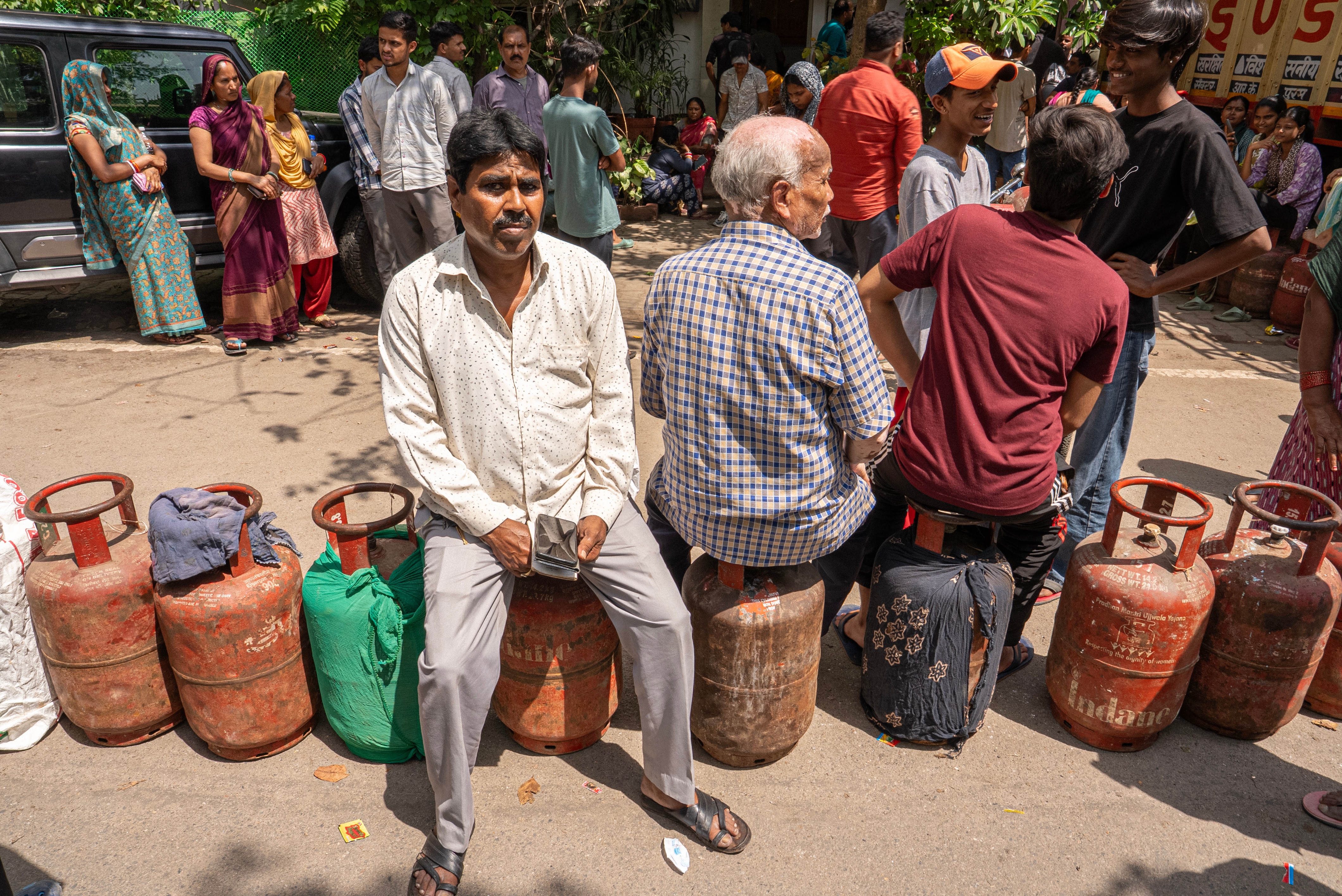Riyasat is serious and sits on a canister as he waits for cooking gas. Other people are sitting with back to the camera.