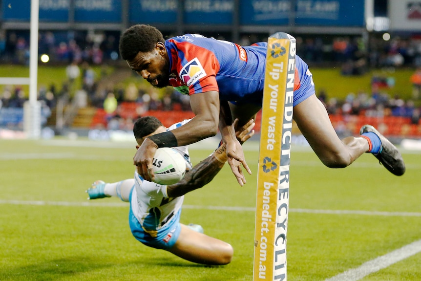 A Newcastle NRL player contorts his body in mid-air to avoid a defender and score a try in the corner.