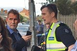 Philip Whiteman on a suburban street holding a phone, standing opposite a police officer.