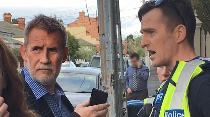 Philip Whiteman on a suburban street holding a phone, standing opposite a police officer.