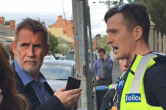 Phillip Whiteman on a suburban street holding a phone, standing opposite a police officer.