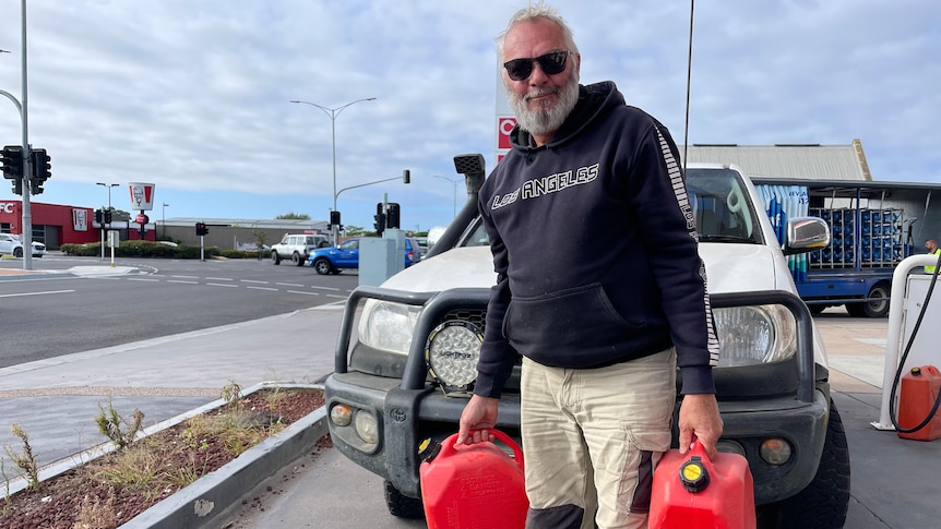 An older, bearded man in dark glasses holds two jerrycans of fuel at a petrol station.