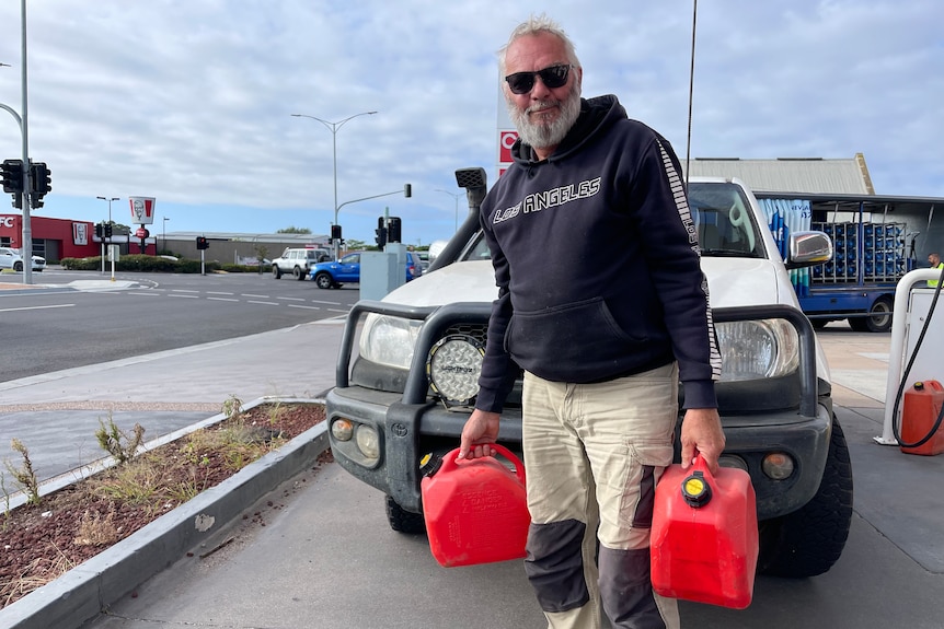 Tony is standing in front of his car holding two red jerry cans at a petrol station in Sale