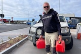 An older, bearded man in dark glasses holds two jerrycans of fuel at a petrol station.