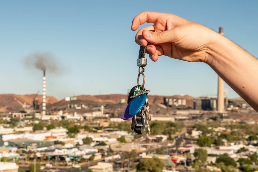 A male hand holds a set of keys in front of the Mount Isa skyline. The town and mine is in the background.