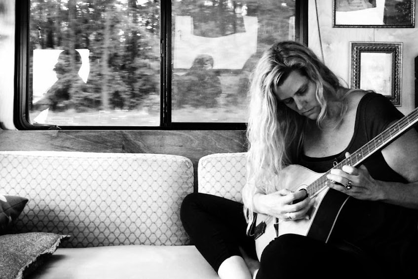Jana sits cross-legged on a couch in the bus playing mandolin in this black and white image taken while the bus is moving.
