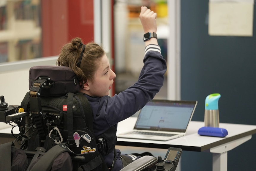 A white child raises his hand in a classroom. He is sitting in a wheelchair in front of a laptop