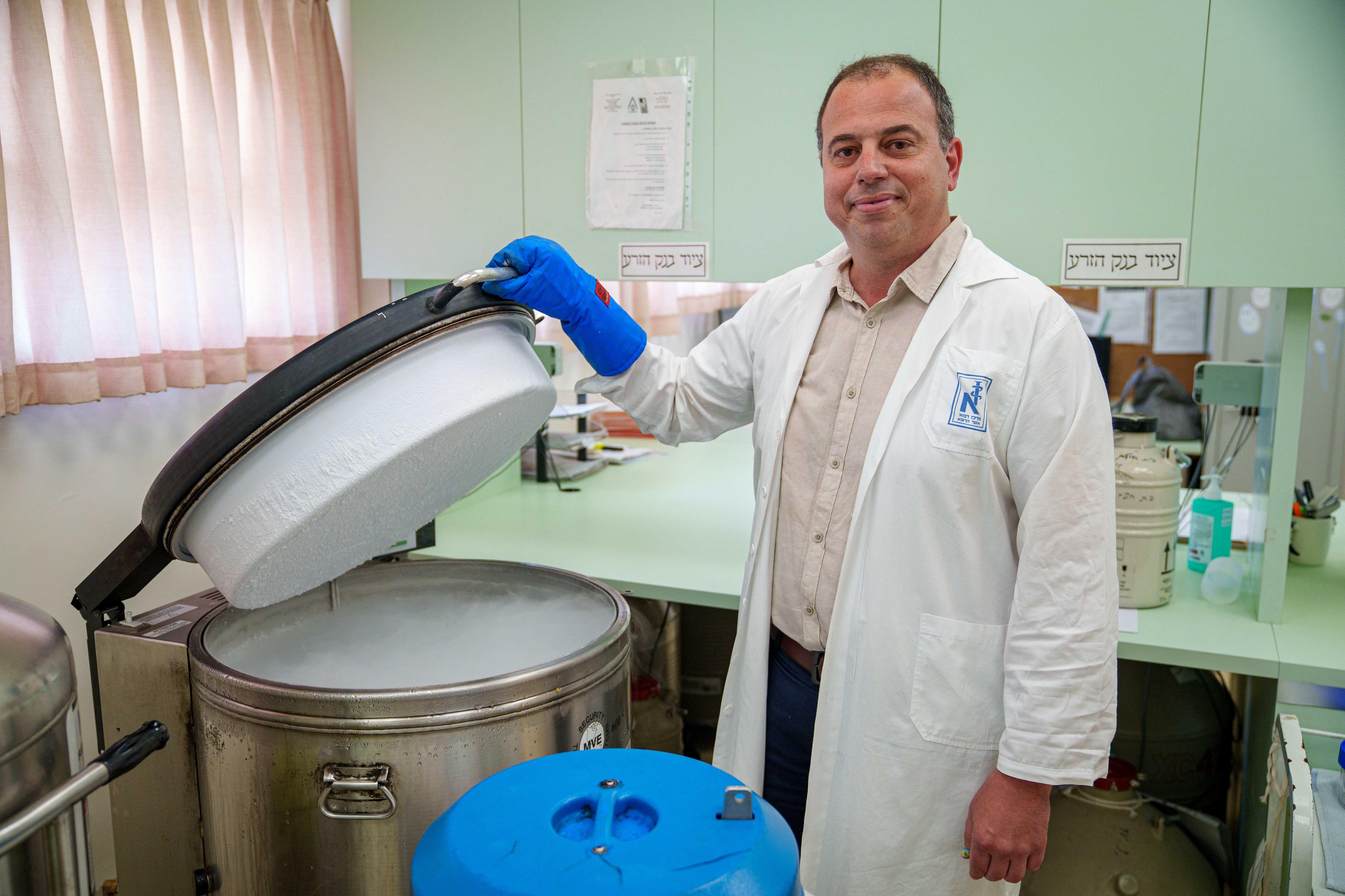 A man wearing a white coat and blue gloves poses while lifting the lid of a machine.