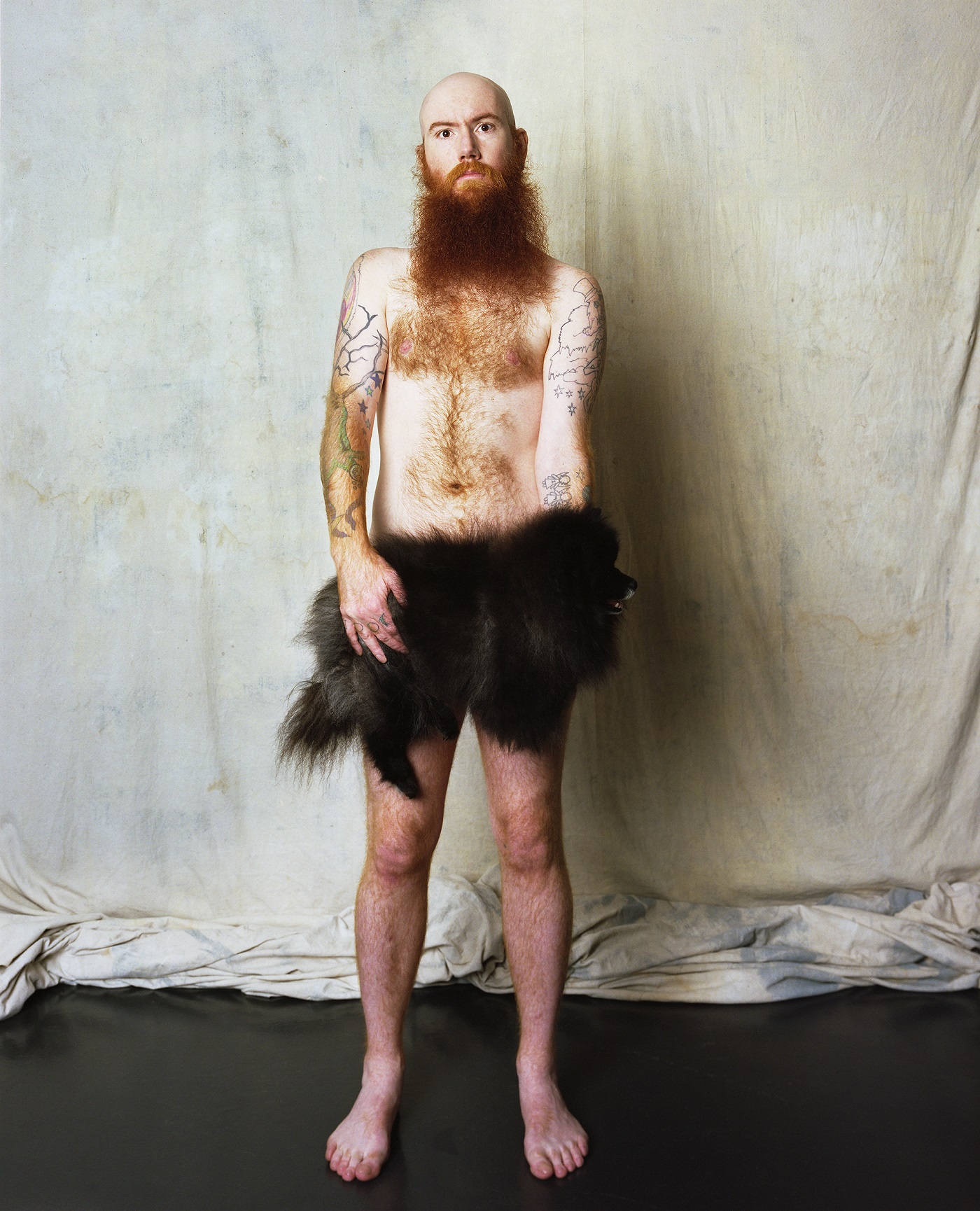 A naked, bearded man holds a fluffy dog.